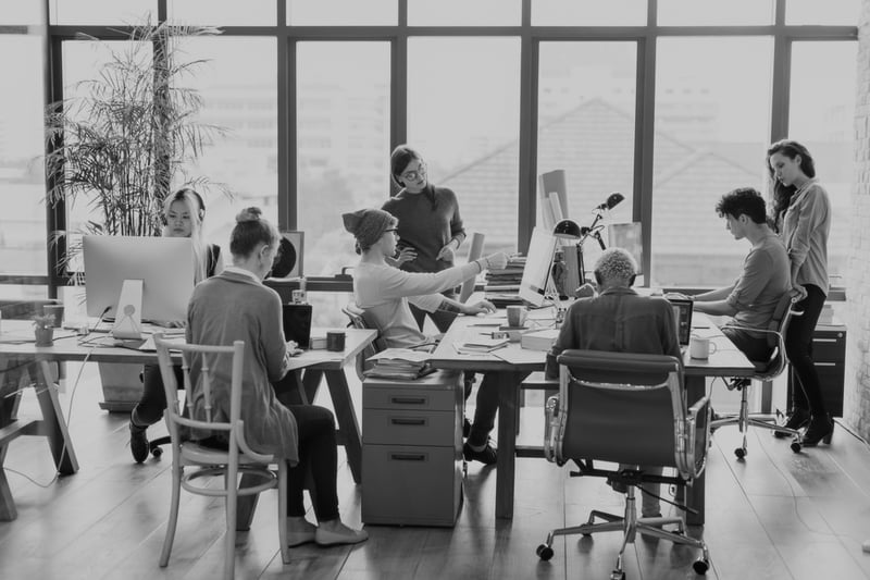 marketing team meeting at desks in large room with windows and desktop computers