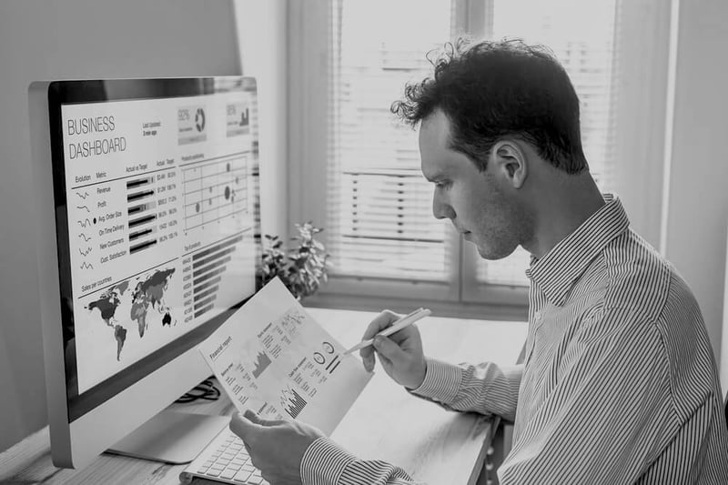 man at desk holding paper with website data in front of computer screen with a business dashboard and analytics