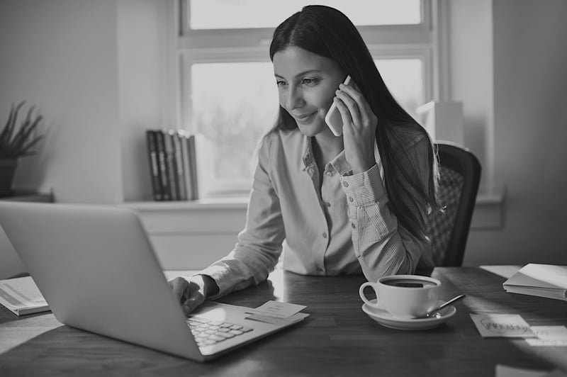 woman on mobile phone and laptop working from home at desk by a window