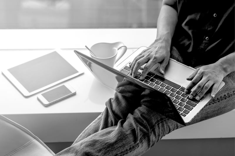 person sitting on windowsill using laptop on lap with phone and tablet beside them